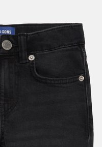 Jean en denim noir avec un design à cinq poches ; comporte des boutons en métal argenté et des coutures ; la ceinture affiche une étiquette bleue avec le nom de la marque.