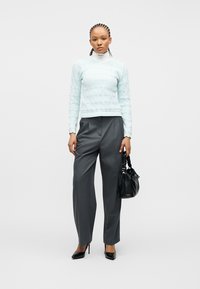 Femme aux cheveux tressés portant un pull en tricot bleu clair sur un col roulé blanc, un pantalon gris, des talons noirs, et tenant un sac à main noir.