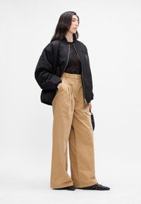 Sort bomberjakke, gennemsigtig sort top, beige brede bukser og sorte spidse sko. Funktioner inkluderer lynlås-lukning og sidelommer.