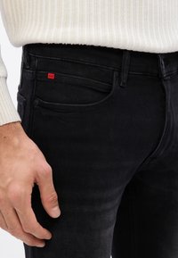 Jeans de mezclilla negra con corte slim, bolsillo frontal visible y etiqueta de logo roja. La tela tiene una ligera textura y decoloración para un aspecto desgastado.