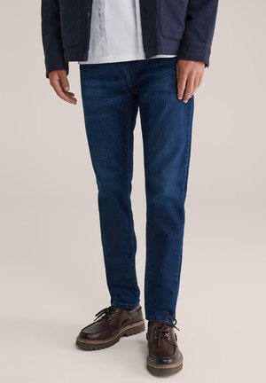 Dunkelblaue Jeans im Slim-Fit-Stil aus Denim, ausgestattet mit einem strukturierten Stoff und subtilen Ausbleichungen. Kombiniert mit dunkelbraunen Schnürschuhen.