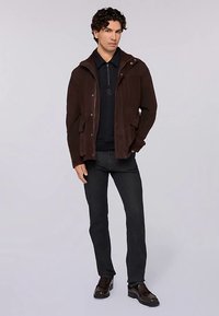 Brun jacka med dragkedja och huva, två framfickor, ovanpå en svart pikétröja, kombinerad med mörka jeans och bruna skor.