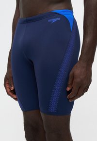 Близък план на човек, носещ тъмносини шорти за плуване Speedo с светлосини зигзагообразни шарки отстрани и светлосини акценти на колана.