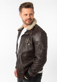 Bruine leren jas met een schapenvachtkraag, gedetailleerd met patches, een ritszak op de arm en geribbelde manchetten. Gekenmerkt over een wit shirt.