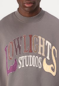 Szürke pulóver, különböző színekben hímzett "LOWLIGHTS STUDIOS" felirattal, bordázott nyakrésszel.