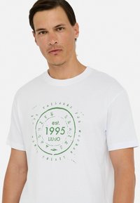 Baltos medvilnės marškinėliai su žalia apvalia grafika, kurioje yra tekstas „est. 1995“ ir „LIU-JO“. Trumpomis rankovėmis ir klasikinio apvalaus kaklo dizainas.