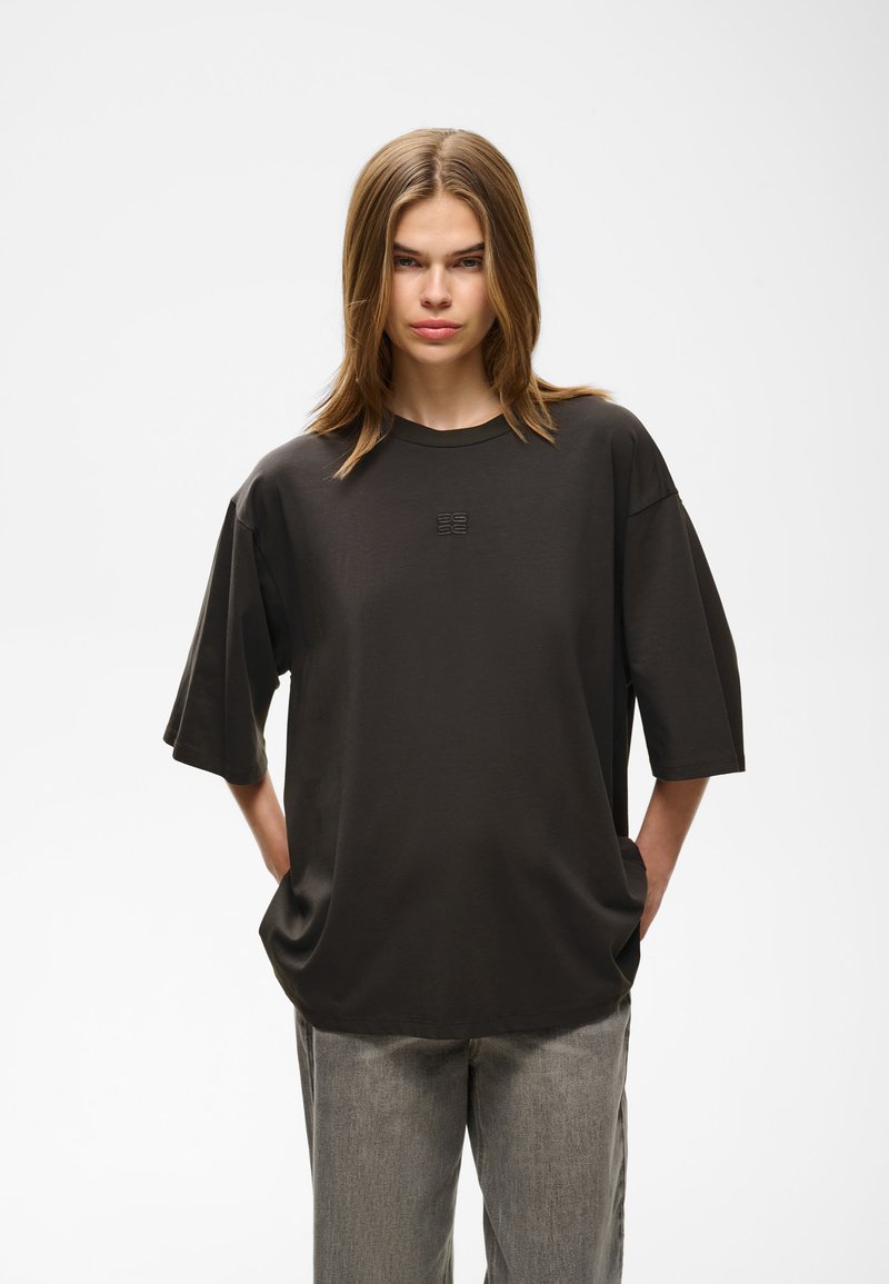 Jeune femme aux cheveux bruns mi-longs porte un t-shirt noir ample et un pantalon gris, debout les mains dans les poches devant un fond uni.
