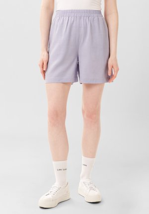 Shorts lavande clair avec une taille élastique, fabriqués à partir d'un tissu doux. Comprend des poches latérales et une coupe décontractée. Portés avec des chaussettes blanches à la cheville et des baskets.