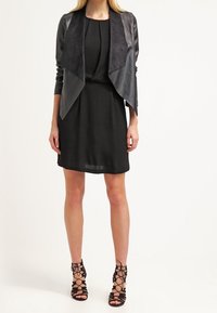 Robe noire portefeuille avec une veste en suède et en cuir, et des escarpins noirs à lanières. La veste a un devant drapé et des manches ajustées.