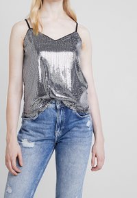 Femme portant un haut camisole à sequins argentés métalliques avec des fines bretelles, associé à un jean déchiré bleu clair devant un fond uni.