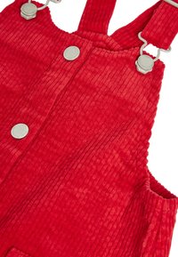 Salopette en velours côtelé rouge avec bretelles réglables, fermoirs en métal, grands boutons argentés et tissu côtelé texturé. Elle possède deux poches latérales.