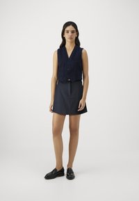 Marineblå vest med en cropped design, knapper og en frontlomme; kombinert med en mørk skaterkjørt og svarte loafers.