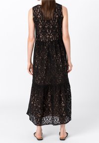 Černé krajkové maxi šaty s vrstveným designem, bez rukávů, s vroubkovaným lemem a složitými květinovými vzory po celém povrchu. K šatům jsou obuty sandále.