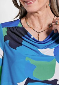 Blaue und grüne blumige Bluse mit gerafftem Ausschnitt und glattem Stoff. Accessoires umfassen eine Mehrfarbige Perlenkette, Ohrringe und Ringe.