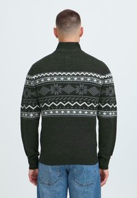 Sötétzöld kötött pulóver magas gallérral, amely szürke és fehér geometrikus mintával díszített a mellkason és az ujjakon.