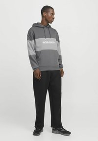 Grau farblich abgesetzter Hoodie mit einem weißen Logo, ausgestattet mit eine Kapuze mit Zugband. Kombiniert mit schwarzen Hosen und schwarzen Sneakers. Glatte Textur.