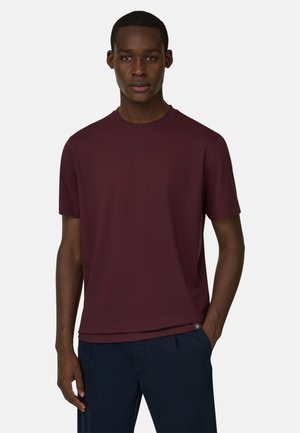 Ung mand iført en ensfarvet vinrød kortærmet t-shirt og mørkeblå bukser, stående med den ene hånd i lommen mod en hvid baggrund.