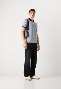 Lys grå polo skjorte med sort krave, sorte jeans og hvite sneakers. Modellen bærer en liten grå skulderveske. Enkelt, moderne design.