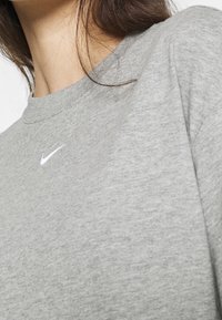Zbliżenie osoby w szarej bluzie Nike z okrągłym dekoltem, z białym logo swoosh umieszczonym na środku klatki piersiowej.