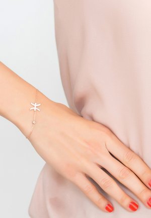 Gold chain bracelet featuring a starfish design with a small gemstone accent, worn on a hand with manicured red nails against a soft pink background.