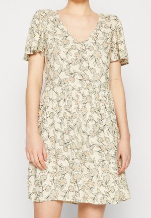 Kobieta w krótkiej kremowej sukience z dekoltem w serek i kwiatowym nadrukiem, stojąca z ramionami swobodnie opuszczonymi wzdłuż ciała na jednolitym tle.