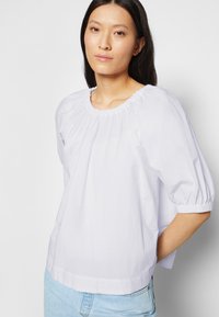 Blusa de algodão branca com mangas bufantes, decote redondo e detalhe franzido. Apresenta um corte solto e textura suave.