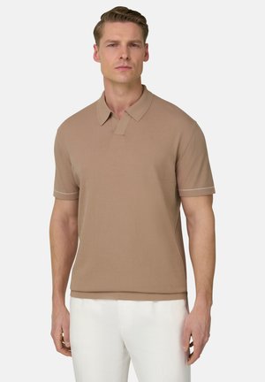 Mann trägt beige Poloshirt mit kurzen Ärmeln und weiße Hose, steht vor weißem Hintergrund, blickt nach vorne mit neutralem Gesichtsausdruck.