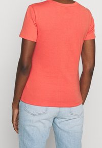 Coral T-shirt with short sleeves and a round neck. Smooth texture; fitted design. Paired with light blue denim jeans, featuring back pockets.