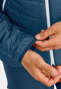 Blaue isolierte Jacke mit gestepptem Textur, weißen Reißverschluss und elastischen Bündchen, während Hände den Ärmel anpassen.