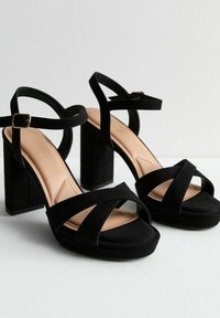 Sandalias negras de tacón alto con gruesos tacones de bloque, tiras de ante entrecruzadas y una plantilla acolchada beige. Cuenta con una correa de tobillo con hebilla para el cierre.