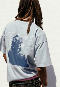 Person med lange dreadlocks står overfor en hvid murstensvæg, iført en lyseblå t-shirt med grafik af en person i VR-headset med teksten "STWD GOALS."