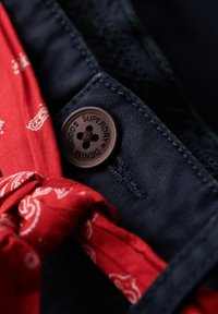 Koszula dżinsowa w ciemnoniebieskim kolorze, ozdobiona czerwoną chustą w wzór paisley związaną przy kołnierzu oraz brązowym guzikiem z detalem logo.