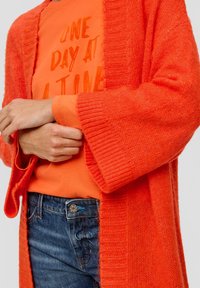Person, der eine orange Strickjacke über einem orangefarbenen Shirt mit dem Text "EIN TAG NACH DEM ANDEREN" trägt, kombiniert mit blauen Jeans.