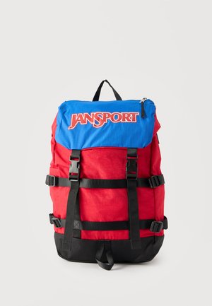 Zaino Jansport rosso e nero con patta blu che presenta un grande logo Jansport, due cinghie nere con fibbie e cinghie laterali regolabili.