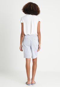 Blouse blanche à manches courtes avec une fente dans le dos, associée à un short mi-long rayé bleu et blanc. Sandales et tatouages visibles sur les bras.