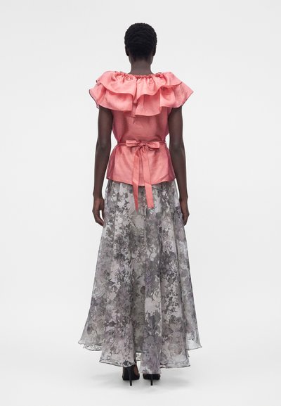 Γυναίκα με ροζ μπλούζα με βολάν δεμένη στη μέση, συνδυασμένη με μια μακριά, διάφανη φλοράλ γκρι φούστα και μαύρες ψηλές γόβες, στεκόμενη με την πλάτη στραμμένη.