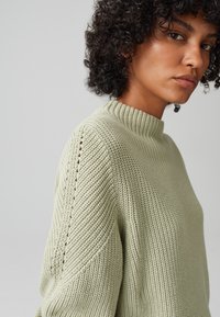 Hellgrüner Strickpullover mit hohem Kragen, strukturierter Musterung und schrägen Rippendetails an den Schultern und Seiten.