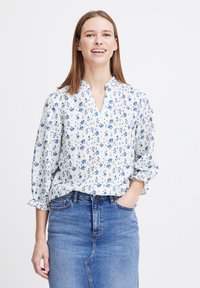 Blouse blanche avec motif floral bleu, décolleté en V et manches 3/4, associée à une jupe en jean. Le tissu semble léger et texturé.