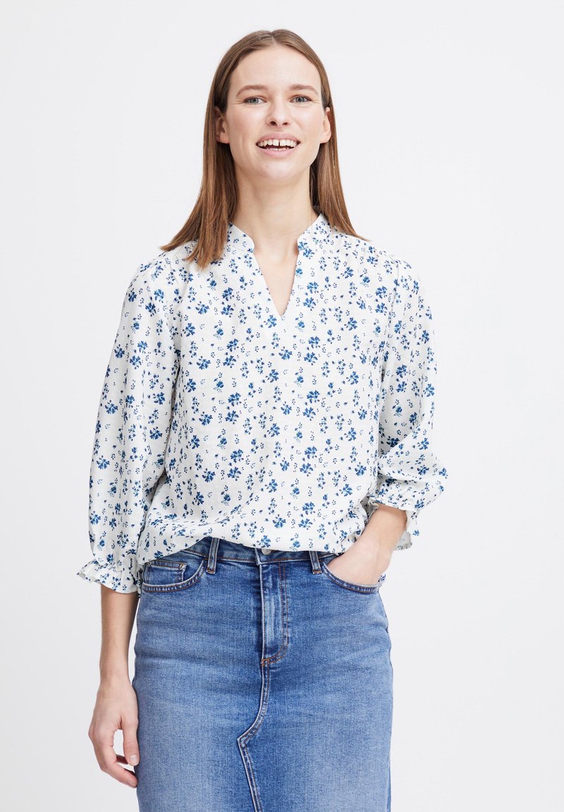 Blouse blanche avec motif floral bleu, décolleté en V et manches 3/4, associée à une jupe en jean. Le tissu semble léger et texturé.