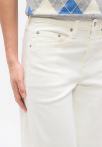 Κοντινό πλάνο ενός ατόμου που φοράει λευκό τζιν και ένα πουλόβερ argyle σε αποχρώσεις του μπ λε, γκρι και εκρού, με το χέρι ακουμπισμένο στο πλάι.