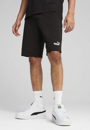 Jambes inférieures d'une personne portant un short Puma noir, des chaussettes blanches Puma et des baskets blanches Puma avec des bandes noires, debout sur un fond neutre.