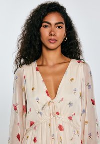 De bloemenjurk heeft een beige achtergrond met rode, blauwe en gele bloemen, een diepe V-hals en een geraffineerde taille met kanten details.