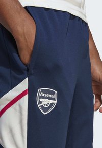 Pantalones cortos deportivos en azul marino con un acabado texturizado, que presentan un logo blanco de Arsenal y detalles rojos en el lateral. Bolsillos integrados en el diseño.