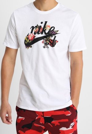 T-shirt blanc Nike avec logo floral noir associé à un short de camouflage Nike rouge, noir et blanc porté par une personne.