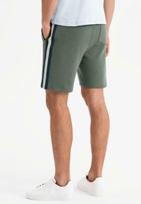 Pantaloni sportivi verdi con strisce laterali blu navy, tasche laterali e un design aderente, realizzati in tessuto morbido, abbinati a sneakers bianche.