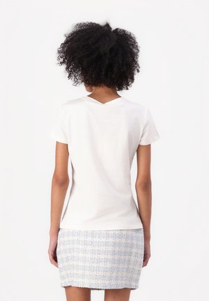 Kobieta z kręconymi włosami, ubrana w gładką białą koszulkę z krótkim rękawem oraz jasnoniebieską teksturowaną mini spódnicę, zwrócona tyłem do aparatu.