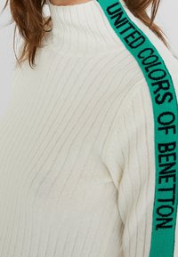 Suéter de cuello alto de color crema con un acabado texturizado y una franja de logo en verde y negro a lo largo del hombro.