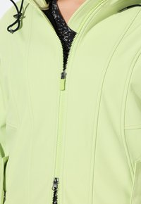 Veste verte claire en tissu lisse, avec un col haut, une fermeture éclair frontale entière et des accents noirs. Elle dispose de poches dissimulées et de détails cousus.