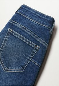 Jean denim bleu plié montrant la poche arrière et la ceinture avec des coutures visibles sur un fond clair.