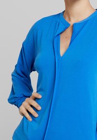 Blå blus med lång ärm i mjukt tyg, med V-halsringning och framplacket. Drapering vid ärmsluten.